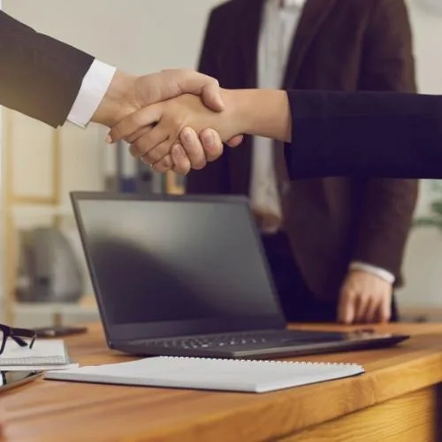 Dos personas se dan la mano sobre un escritorio con una computadora portátil